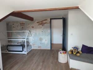 Chambre enfant mansardée : lit cabane, sol bois, poutres foncées. Papier peint carte du monde d'aventure, jouets et pouf de rangement.