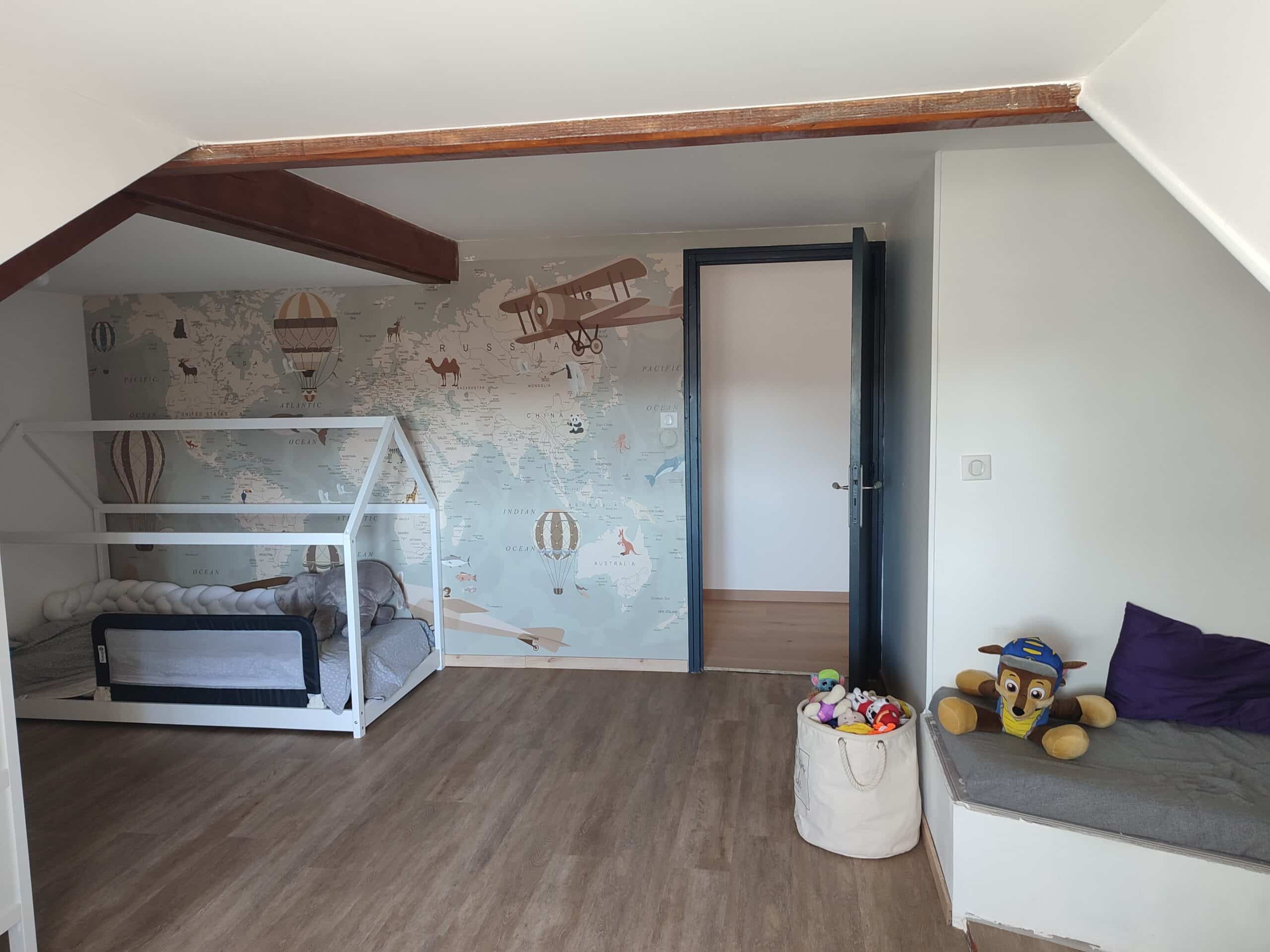 Chambre enfant mansardée : lit cabane, sol bois, poutres foncées. Papier peint carte du monde d'aventure, jouets et pouf de rangement.