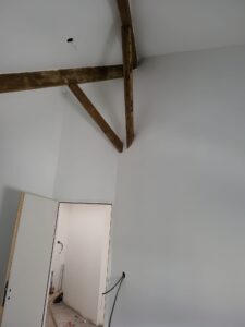 Intérieur moderne en rénovation. Murs gris clair contrastant avec des poutres de charpente en bois rustiques. Porte ouverte, câbles électriques visibles.